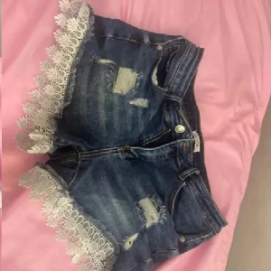 Blå jeansshorts med spetskant - Snygga blå jeansshorts med slitna detaljer och coola hål framtill. Kanterna är dekorerade med vit spets som ger en extra touch. Klassisk femficksmodell med knapp och dragkedja. Perfekta för dig som vill sticka ut lite extra i sommar.