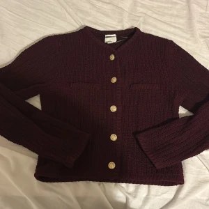 Vinröd stickad kofta från H&M - Mysig vinröd stickad kofta från H&M med snygga guldknappar framtill och två dekorativa fickor på bröstet. Koftan har långärmad passform och är perfekt för lager-på-lager under kyliga dagar.