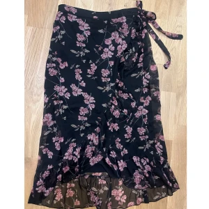 Svart blommig omlottkjol - Svart omlottkjol med rosa blommor och volangkant. Kjolen har ett band i midjan man kan knyta. Använd två gånger. Storlek M/38.