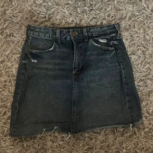 Blå jeanskjol från &Denim - Säljer en blå jeanskjol från &Denim i klassisk femficksmodell med rå kant nedtill och slitna detaljer. Kjolen har knapp och dragkedja framtill samt bakfickor. Perfekt för dig som gillar en chill och edgy stil.