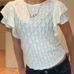 Vit blus med volangärmar - Säljer en vit blus med snyggt rutmönster och volangdetaljer på ärmarna. Blusen har rund hals och är kortärmad, perfekt för dig som gillar en fräsch och feminin stil. Materialet är lätt och luftigt, vilket gör den skön att bära under varma dagar.