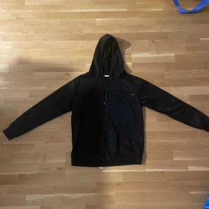 Svart hoodie med tryck Eat Sleep Rave Repeat - Svart hoodie med huva och dragkedja framtill. På ryggen finns ett stort vitt tryck med texten 'Eat Sleep Rave Repeat' och tillhörande ikoner. Tröjan har långa ärmar, snörning vid huvan och en klassisk känguruficka. Perfekt för dig som gillar rave och streetstyle.