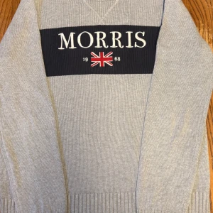 Grå stickad tröja från Morris - Snygg grå stickad tröja från Morris med mörkblå panel och broderad logga samt brittisk flagga framtill. Tröjan har rund halsringning, ribbade muddar och lång ärm. Perfekt för dig som gillar klassisk och stilren look.