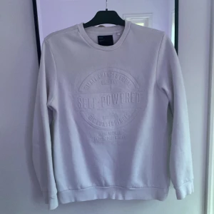 Vit sweatshirt från Bershka - Vit sweatshirt från Bershka med upphöjt tryck på bröstet där det står 'SELF-POWERED'. Tröjan har rund halsringning, långa ärmar och ribbade muddar. Perfekt för en chill och stilren look.