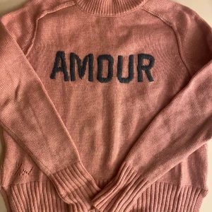 Rosa stickad tröja med text Amour - Säljer en stickad rosa tröja med svart texten 'AMOUR' framtill. Tröjan har ribbade muddar vid ärmslut och nederkant samt lång ärm. Perfekt för dig som vill ha en mysig och trendig look. BARN STORLEK 12 år Nypris 1500 köpt på NK i Stockholm