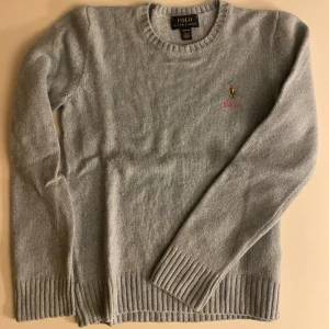 Ljusgrå stickad tröja från Polo Ralph Lauren - Snygg ljusgrå stickad tröja från Polo Ralph Lauren med klassisk rund halsringning och broderad logga på bröstet. Tröjan har långa ärmar och ribbade muddar vid ärmslut och nederkant. Perfekt inför Hösten!!! BARN STORLEK