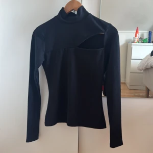 Svart polotopp med cut-out från NA-KD - Svart långärmad polotopp från NA-KD i storlek XS. Toppen har en cool asymmetrisk cut-out vid axeln och är figurnära i modellen. Materialet är mjukt och stretchigt, troligtvis polyester. Perfekt för dig som gillar minimalistisk stil med en twist.