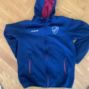 Blå hoodiejacka CCM DIF strl L - Snygg blå hoodiejacka från CCM med Djurgårdens IF-logga broderad på bröstet. Jackan har dragkedja, två fickor med röda detaljer och justerbar huva med röd insida. Perfekt för dig som vill visa DIF-stolthet och hålla dig varm.