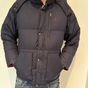 Mörkblå Ralph lauren puffer-jacket S/M - Snygg och varm dunjacka i mörkblå färg med röd insida och klassisk Polo-logga broderad på bröstet. Jackan har en sportig men stilren design, perfekt för kalla höst- och vinterdagar