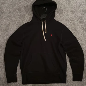 Svart Polo Ralph Lauren hoodie - Svart hoodie från Polo Ralph Lauren med klassisk röd logga broderad på bröstet. Tröjan har huva med vita snören och en stor magficka framtill. Perfekt för dig som gillar stilren och enkel design. Pris kan diskuteras!