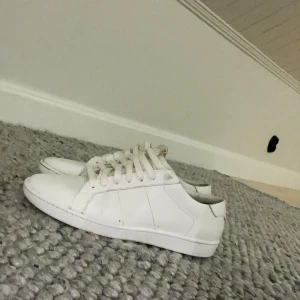 Vita sneakers från Saint Laurent - Säljer ett par stilrena vita sneakers från Saint Laurent. Skorna har klassisk låg profil, rund tå och snörning framtill. Tillverkade i vitt skinn med diskret branding på hälen. Perfekta för en clean och minimalistisk look. De ör model al/01 de är i skick 9 av 10 priser är inte hugget i sten. Storlek 39,5 sitter som 41 ungefär 