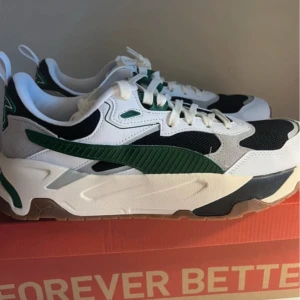 Puma sneakers i vitt, grönt och svart - Snygga Puma sneakers med chunky sula i vitt, grönt och svart. Skorna har klassisk snörning, gröna detaljer och logga baktill. Ovandelen är i mesh och syntet med coola kontraster och modern streetstil. Perfekta för dig som vill sticka ut med en sportig vibe.