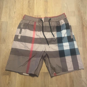 Burberry shorts - Snygga badshorts från Burberry med klassiskt rutigt mönster i beige, svart, röd och blå. Shortsen har elastisk midja med svart snörning och praktiska fickor fram och bak. Perfekta för sommaren och strandhäng!