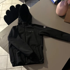 Svart vindjacka från Sail Racing - Svart vindjacka från Sail Racing med huva och dragkedja framtill. Jackan har två fickor med dragkedja och är fodrad med mjuk fleece på insidan. Perfekt för blåsiga dagar och snygg, stilren look.