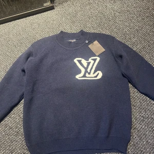 stickad tröja Louis Vuitton - Exklusiv mörkblå stickad tröja från Louis Vuitton med stort vitt LV-logo på både fram- och baksidan. Tröjan har rund hals, ribbade muddar och är långärmad. Perfekt för dig som vill ha en lyxig och stilren look.