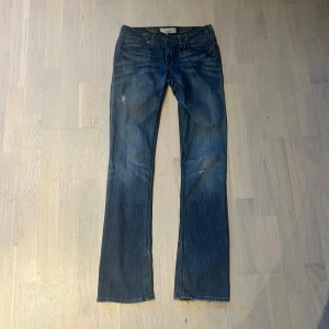 True Religion blå bootcut jeans - Snygga blå jeans från True Religion med bootcut passform och broderade bakfickor. Jeansen har klassisk femficksdesign, låg midja och slitna detaljer för en cool look. Tillverkade i jeansmaterial med tydliga kontrastsömmar och dekorativa broderier.
