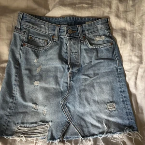 Ljusblå jeanskjol från &Denim - Säljer en kort ljusblå jeanskjol från &Denim med slitna detaljer och fransad nederkant. Kjolen har klassiska fickor fram och bak🤍