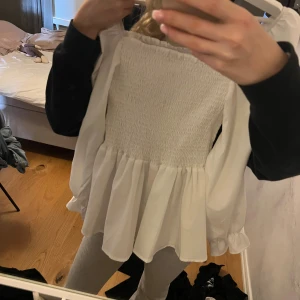 Vit blus med volang och smock - Säljer en vit blus med smockad överdel och volang nedtill. Blusen har långa ärmar med lätt puff och resår vid ärmslut. Perfekt för dig som gillar romantisk och söt stil. Passar till jeans eller kjol för en trendig look.