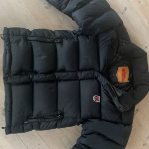 Svart dunjacka från Fjällräven XXS - Säljer en svart, puffig dunjacka från Fjällräven i storlek XXS. Jackan har klassisk Fjällräven-logga på bröstet, två dragkedjeförsedda fickor vid sidan. Tyvärr har jackan ett litet hål på ryggen längst ned (se sista bilden)