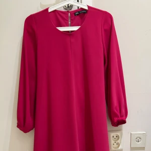 Ceriserosa klänning från Zara - Superfin ceriserosa klänning från Zara med rund halsringning och långa ärmar. Klänningen har ett knytband i midjan och stängs med knapp i nacken. Den är lätt utställd och slutar ovanför knäna, perfekt för dig som gillar färg och enkel stil.