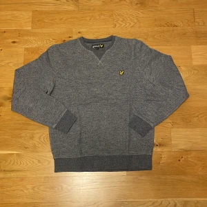 Grå sweatshirt från Lyle & Scott - Tjena säljer en gråmelerad sweatshirt från Lyle & Scott med klassisk rund hals och broderad gul logga på bröstet. Tröjan har ribbade muddar vid ärmslut och nederkant, perfekt för en chill och stilren look.