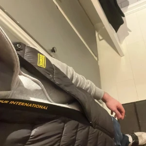 Svart dunväst från Barbour International - Säljer en svart dunväst från Barbour International med gul logotyp på bröstet och dragkedja med märkets namn. Västen är quiltad och har en hög krage. Perfekt att slänga över en hoodie för en cool och sportig look.