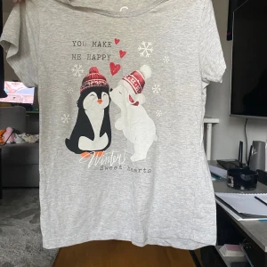 Grå t-shirt med vintermotiv - Söt grå t-shirt med tryck av en pingvin och en isbjörn i mössa, snöflingor och röda hjärtan. Texten 'You make me happy' och 'Winter sweet hearts' på framsidan. Perfekt för dig som gillar gulliga vintermotiv och mjuka material.