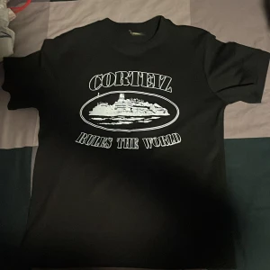 Svart Corteiz t-shirt med tryck - Svart t-shirt från Corteiz i storlek small med stort vitt tryck på framsidan där det står 'Corteiz Rules The World' och en grafisk bild. Klassisk rund hals och korta ärmar. Materialet känns som mjuk bomull, perfekt för en chill streetwear-look.