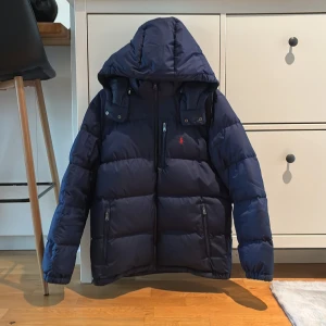 Mörkblå pufferjacka Polo Ralph Lauren XL - Säljer en mörkblå pufferjacka från Polo Ralph Lauren i storlek XL. Jackan har huva, tre dragkedjefickor och klassisk röd broderad logga på bröstet. Perfekt för kalla dagar och riktigt snygg streetstil.