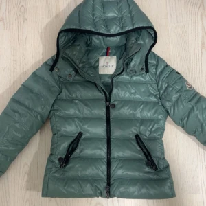 Grön pufferjacka från Moncler - Snygg grön pufferjacka från Moncler med huva och svarta dragkedjor. Jackan har quiltad design, två fickor med dragkedja och klassisk Moncler-logga på ärmen. Perfekt för kyliga dagar och riktigt skön passform.Etiketten är lite lös, bilden är felaktig.