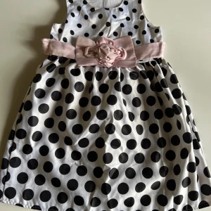 Prickig klänning med rosett från To Sleep Bear - Supersöt vit klänning med stora svarta prickar och bred ljusrosa rosett i midjan. Klänningen är ärmlös och har en dekorativ blomdetalj framtill. Tillverkad i glansigt material som ger en festlig känsla. Dragkedja i ryggen för enkel påklädning.