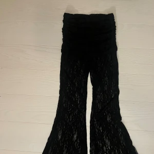 Svarta utsvängda spetsbyxor - Säljer ett par svarta byxor i transparent spets med blommigt mönster. Byxorna har en utsvängd passform nertill för en riktigt snygg siluett. Perfekta för dig som vill sticka ut med en unik och trendig look.