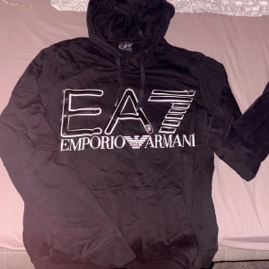 Svart EA7 Emporio Armani hoodie - Svart hoodie från EA7 Emporio Armani med stor vit logotyp på bröstet. Tröjan har huva med snörning och en klassisk magficka. Mjuk bomullskvalitet och långärmad modell. Hoodien är köpt från JD och är i storlek xs. Den sitter bra på både män och kvinnor. Nästan aldrig använd.