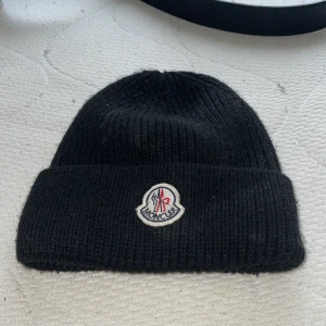 Svart Moncler mössa i ull - Svart ribbstickad mössa från Moncler med klassisk uppvikt kant och broderad logotyp framtill. Snygg och enkel design som passar perfekt till kalla dagar. Tillverkad i mjuk ull för extra värme och komfort.