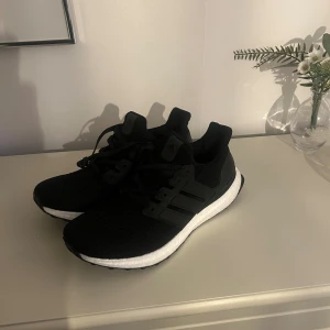 Adidas Ultraboost svarta sneakers - Svarta Adidas Ultraboost sneakers med vit Boost-sula och stickad ovandel. Skorna har klassiska tre ränder på sidan, snörning och en bekväm, responsiv sula. Perfekta för dig som gillar sportig stil och vill ha riktigt sköna skor till vardagen.