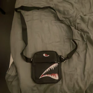Svart axelväska med hajmotiv - Cool svart axelväska med justerbart band och ett unikt hajmotiv på framsidan. Väskan har en fyrkantig form och dragkedja upptill. Perfekt för att bära dina viktigaste grejer och sticka ut med en edgy stil. Helt ny! Pris kan diskuteras!