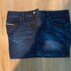 Mörkblå jeanskjol från Diesel - Snygg mörkblå jeanskjol från Diesel med klassiska fickor fram och bak, bälteshällor och knappstängning. Kjolen har en rak, kort modell och är tillverkad i denim med coola slitningar. Perfekt för dig som gillar en avslappnad och trendig stil.