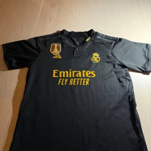 Real Madrid Bellingham svart matchtröja - Snygg svart Real Madrid fotbollströja från Adidas med gul broderad klubbmärke, FIFA World Champions 2022 patch och Emirates Fly Better tryck. Bellingham och nummer 5 på ryggen. Kortärmad med klassisk krage och knapp. Tillverkad i lätt polyester.