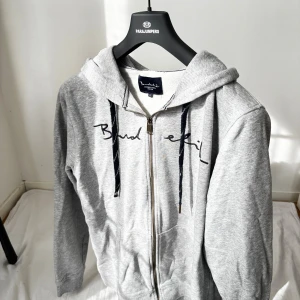 Grå hoodie med dragkedja Bondelid - Grå hoodie från Bondelid med svart broderad logga över bröstet. Tröjan har huva med snörning, hel dragkedja framtill och två fickor. Tillverkad i mjuk bomullsblandning, perfekt för chill dagar. Klassisk och enkel stil med långa ärmar. (storlek L)