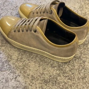 Lanvin sneakers i mocka och skinn - Snygga sneakers från Lanvin i beige mocka med glansig, ljusbrun tå och matchande detaljer runt öppningen. Skorna har snörning och en platt, vit sula. Perfekta för dig som gillar stilrena och exklusiva sneakers med en twist.