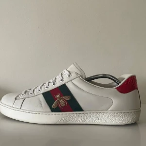 Gucci Ace sneakers med bi-detalj - Gucci Ace sneakers i vitt skinn med klassiska gröna och röda ränder på sidan och broderad guldfärgad bi. Baksidan har metallicröd och metallicgrön hälflik i ormmönstrat material. Snygg och ikonisk modell med snörning och platt sula.