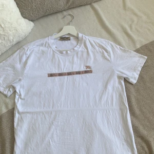 Vit Burberry t-shirt med rutig detalj - Vit t-shirt från Burberry i bomull med klassisk rund hals. På bröstet finns en beige rutig detalj och broderad Burberry-ryttare. Enkel men lyxig design som sticker ut med den ikoniska rutiga randen. Skriv om frågor och pris kan diskuteras👍