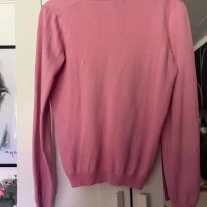 Rosa v-ringad tröja från Gant XS - Snygg rosa tröja från Gant i v-ringad modell. Tröjan är i tunn, stickad bomull och har långa ärmar samt ribbade muddar. Perfekt för dig som gillar klassisk och stilren look med en touch av färg. Liten Gant-logga broderad på bröstet. Tröjan är ganska stor i storlek liknar S. Har några märken på framsidan som inte går bort. 
