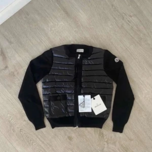 Svart Moncler kofta med quiltad front - Svart kofta från Moncler med quiltad, glansig front och två fickor med knapp. Ärmar och rygg i mjuk stickad kvalitet, dragkedja framtill och Moncler-logga på ärmen. Perfekt för lager-på-lager under höst och vinter.