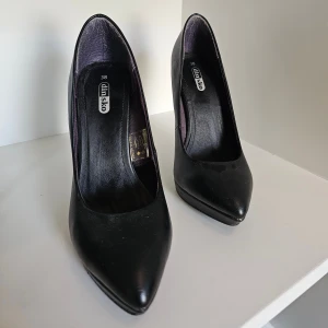 Svarta pumps från Din Sko, storlek 38 - Klassiska svarta pumps från Din Sko med spetsig tå och hög klack. Skorna är tillverkade i syntetmaterial med en stilren, matt finish. Perfekta för dig som vill ha en tidlös och elegant look med extra höjd.