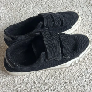 Even odd skor - Svarta sneakers i manchestertyg med vit sula och tre breda kardborreband. Skorna har en klassisk rund tå och platt sula, vilket gör dem både bekväma och stilrena. Perfekta för dig som gillar en enkel men trendig look.