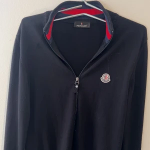 Svart Moncler kofta med dragkedja - Svart kofta från Moncler med hel dragkedja, ribbade muddar och krage med röda och marinblå detaljer. Klassisk Moncler-logga på bröstet. Mjuk och skön, perfekt för lager-på-lager. Passformen är normal och ärmarna är långa.