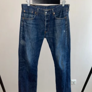 Levi's 501 blå jeans W33 L32 1990-2000 - Klassiska Levi's 501 jeans i blå denim med raka ben och femficksmodell. Jeansen har knäppning fram, tydliga kontrastsömmar och den ikoniska läderpatchen bak i midjan. Perfekta för dig som gillar tidlös stil och vill ha ett par riktigt schyssta jeans.