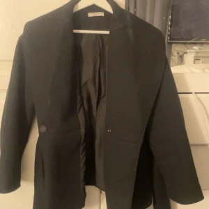 Svart kappa med knytning från Gina Tricot - Säljer en svart kappa från Gina Tricot i storlek XXS. Jackan har ett snyggt bälte i midjan, bred krage och knappar framtill. Materialet känns mjukt och har en klassisk look som funkar till många stilar. Perfekt för dig som gillar stilrena och enkla plagg.