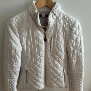 Vit quiltad jacka från Zara - Snygg vit quiltad jacka från Zara med dragkedja framtill och två fickor med guldfärgade dragkedjor. Jackan har ståkrage med knappar och ribbade partier i sidorna för extra komfort. Perfekt för dig som gillar stilren och sportig look